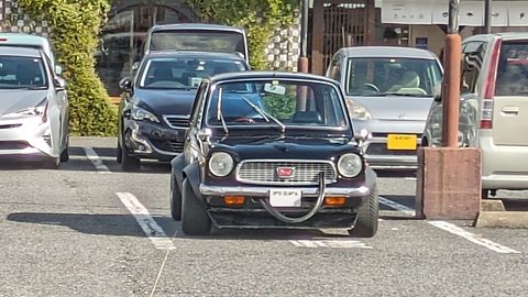 Black small saloon head-on in a car park among modern cars. Opens a larger view