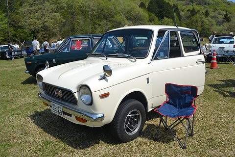 White small saloon on grass with a folding chair beside it and visitors behind. Opens a larger view