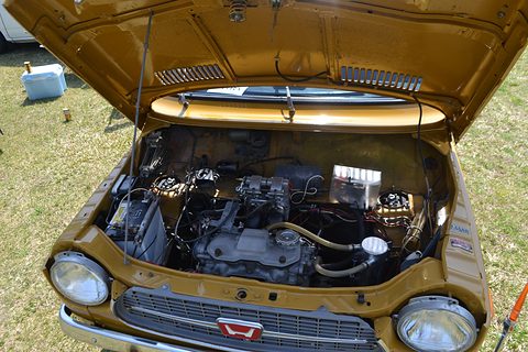 Open bonnet on a mustard-yellow car showing a compact engine with carburettors and wiring. Opens a larger view
