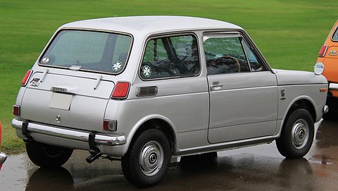 Rear three-quarter of a silver small saloon with a Racing Mate sticker, on wet tarmac. Opens a larger view