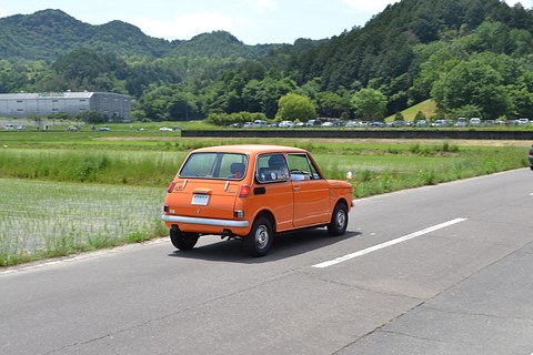 A small orange saloon driving along a road between green fields and wooded hills.