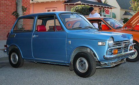 Light blue small saloon with red seats parked by an orange car, a house behind. Opens a larger view