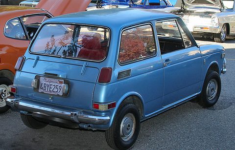 Rear three-quarter view of a small blue two-door saloon with chrome bumpers and red upholstery seen through the glass. Opens a larger view