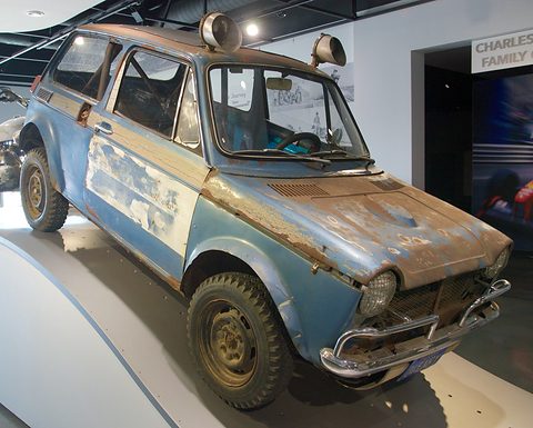 Rusty pale blue small saloon with a roll cage, roof lamps and battered panels on a curved display. Opens a larger view