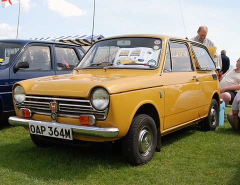 Yellow small saloon on grass with a UK plate, a man behind and a striped awning. Opens a larger view