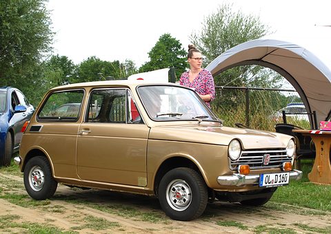 Gold small saloon on grass, a woman standing by a canopy behind it. Opens a larger view