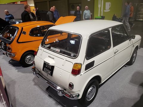 Rear three-quarter of a white small saloon with a dealer sticker, an orange coupé beside it. Opens a larger view