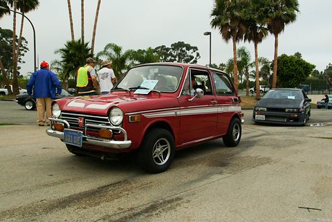 Red small saloon with white stripes driving across a car park lined with palm trees. Opens a larger view