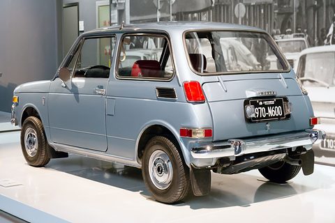 Rear three-quarter of a pale blue small saloon on a white plinth. Opens a larger view