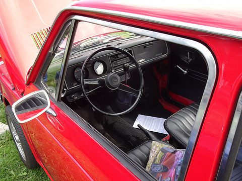 Through an open red door: a dished steering wheel, round dials and black seats. Opens a larger view