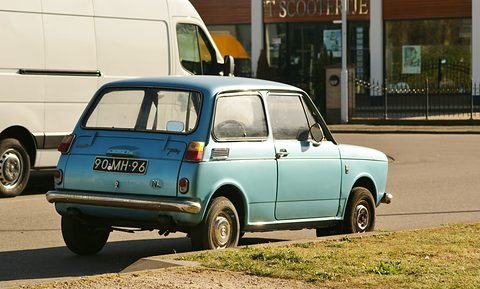Rear three-quarter view of a small pale blue two-door saloon parked by a grass verge, a white van behind. Opens a larger view