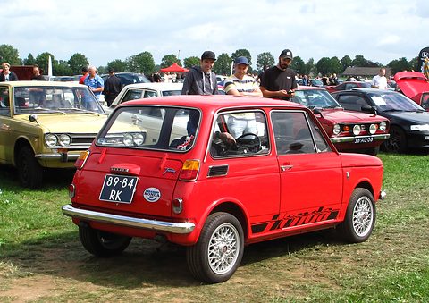 Rear three-quarter of a red small saloon with alloy wheels on grass, men standing behind. Opens a larger view