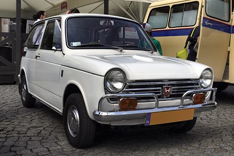 White small saloon with fog lamps on cobbles, a cream bus behind. Opens a larger view