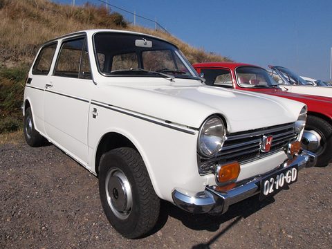 Low front view of a white small saloon on a grassy dune edge, cars behind. Opens a larger view