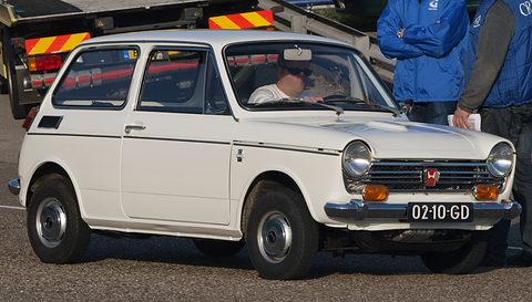 White small saloon with a Dutch plate, its driver seated inside and people standing behind. Opens a larger view