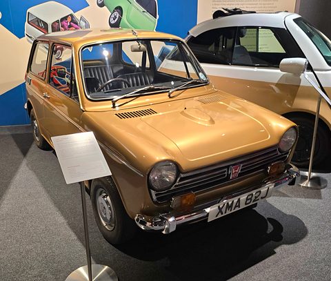 Gold small saloon with a UK plate in front of a bright blue wall with a placard. Opens a larger view