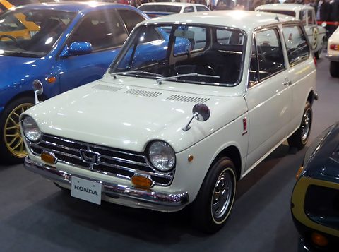 White small saloon on a dark show floor beside a blue sports car. Opens a larger view