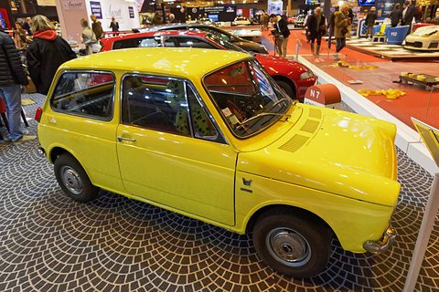 Yellow small saloon on a cobble-patterned show floor in a crowded hall. Opens a larger view