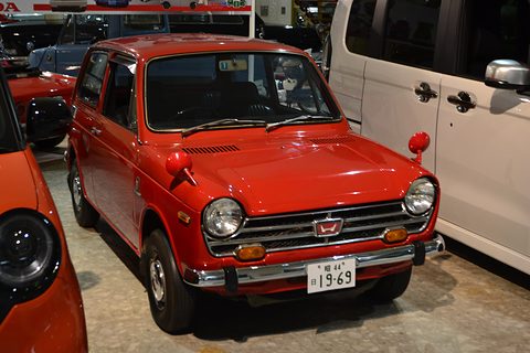 Red small saloon with a 19-69 plate between other cars on a museum floor. Opens a larger view
