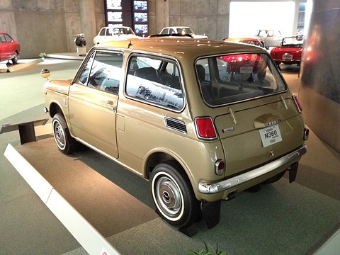 Rear three-quarter of a gold small saloon on a white display base with a placard. Opens a larger view