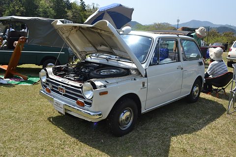 White small saloon with the bonnet raised on grass, its owner sitting under a sunshade beside it. Opens a larger view