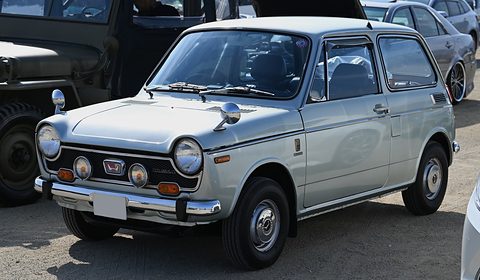 Silver small saloon with twin driving lamps under its grille, an olive truck beside it. Opens a larger view