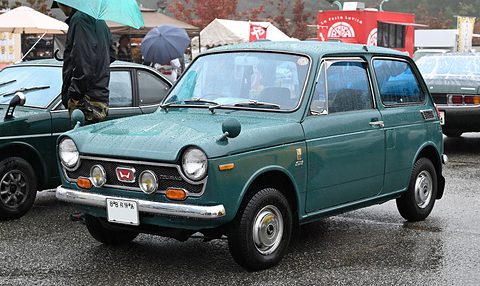 Dark green small saloon with driving lamps on wet tarmac, umbrellas behind. Opens a larger view