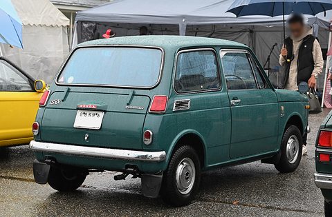 Rear three-quarter of a dark green small saloon with an S badge, a man with an umbrella behind. Opens a larger view