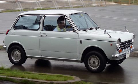 Side view of a pale grey small saloon driving on a wet road, its driver visible inside. Opens a larger view