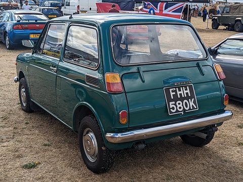 Rear three-quarter of a teal green small saloon on dry grass, vans and flags behind. Opens a larger view