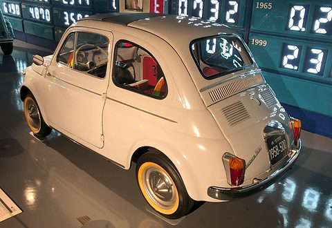 Cream Fiat 500 from the rear right with a louvred engine lid and black plate, on a glossy floor. Opens a larger view