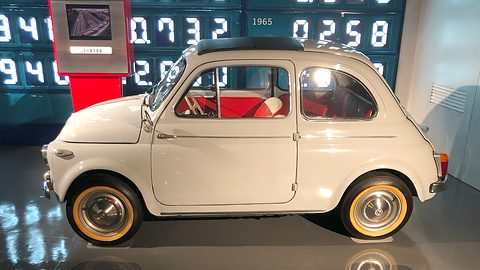Side view of a cream Fiat 500 with red seats and cream wheels on a reflective floor, a digital display behind.