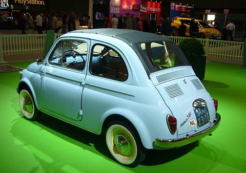Pale blue Fiat 500 from the rear right, its full-length fabric roof and cream wheels, on bright green show carpet. Opens a larger view