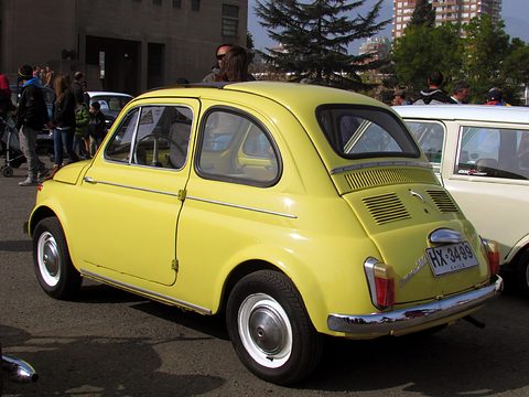Yellow Fiat 500 from the rear left with a louvred engine lid and Chilean plate, a white car and visitors beside it. Opens a larger view
