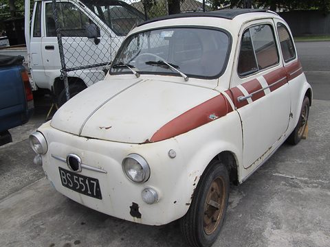 A small white two-door Fiat with a red stripe along its flank, rust on its wing and faded paint, by a chain-link fence. Opens a larger view