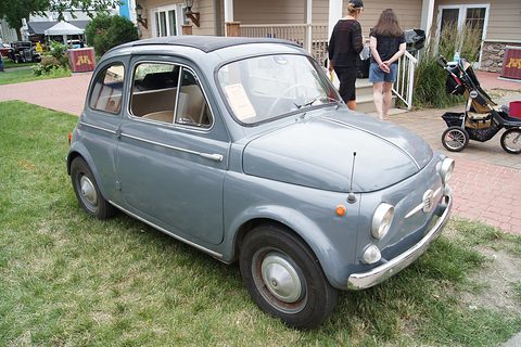 Grey Fiat 500 with a folded fabric roof parked on grass, two people walking past a house behind. Opens a larger view