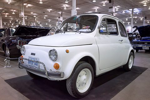 White Fiat 500 with tubular bumper guards and white steel wheels, on black carpet in a large car-show hall. Opens a larger view