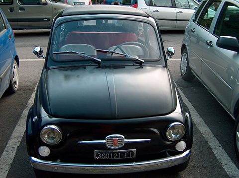 Front of a black Fiat 500 with a chrome grille badge and round headlamps, parked between modern cars. Opens a larger view