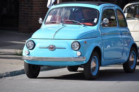 Light blue Fiat 500 with red seats and chrome trim, parked at a kerb before a brick building. Opens a larger view