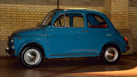 Side view of a blue Fiat 500 with chrome hubcaps, lit against a pale brick wall. Opens a larger view