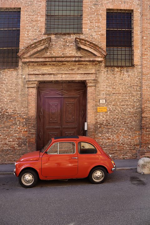 Small red Fiat 500 in profile at the foot of a tall red-brick church front with a carved wooden door. Opens a larger view