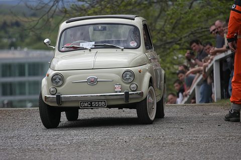 Cream Fiat 500 driving up a steep track towards the camera, spectators leaning on a railing to the right.