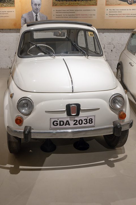 Front of a white Fiat 500 with round headlamps and a red Fiat badge, parked under museum display boards. Opens a larger view