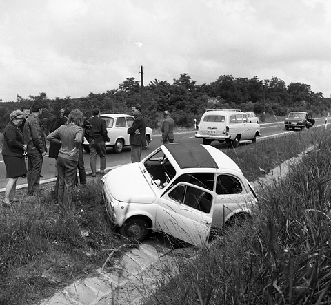 A small white car with its fabric roof open tipped into a grassy ditch, a crowd watching from the roadside. Opens a larger view