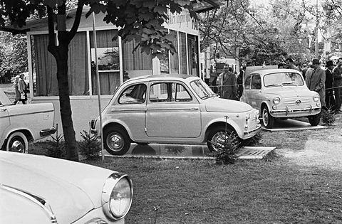 A small pale two-door car in profile on a grass display stand, a larger Fiat and visitors in hats behind. Opens a larger view