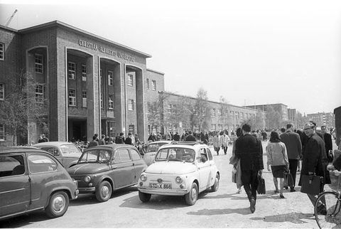 A small white car among parked saloons in front of a brick university building, students walking past. Opens a larger view
