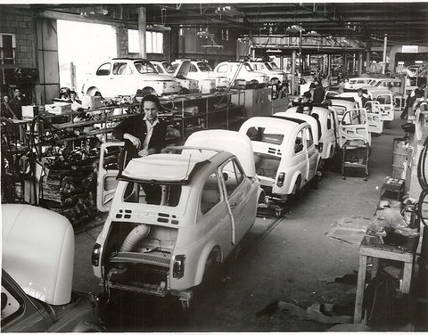 A factory line of white small-car bodies with open engine bays, workers among them and a row of finished cars beyond. Opens a larger view