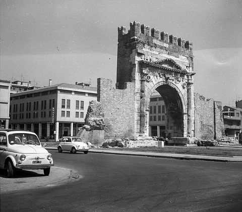 A small white car on a curving road in the foreground, a Roman triumphal arch and modern blocks behind.