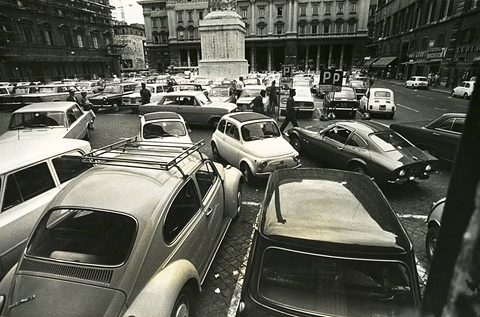 A square crammed with parked and moving cars, several small rounded Fiats among them, a column and palazzi beyond.