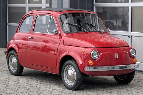Red Fiat 500 with chrome bumpers and hubcaps, parked on grey paving before a glass-fronted building. Opens a larger view